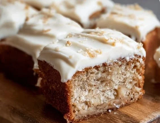 Moist banana cake topped with cream cheese frosting on a white plate.